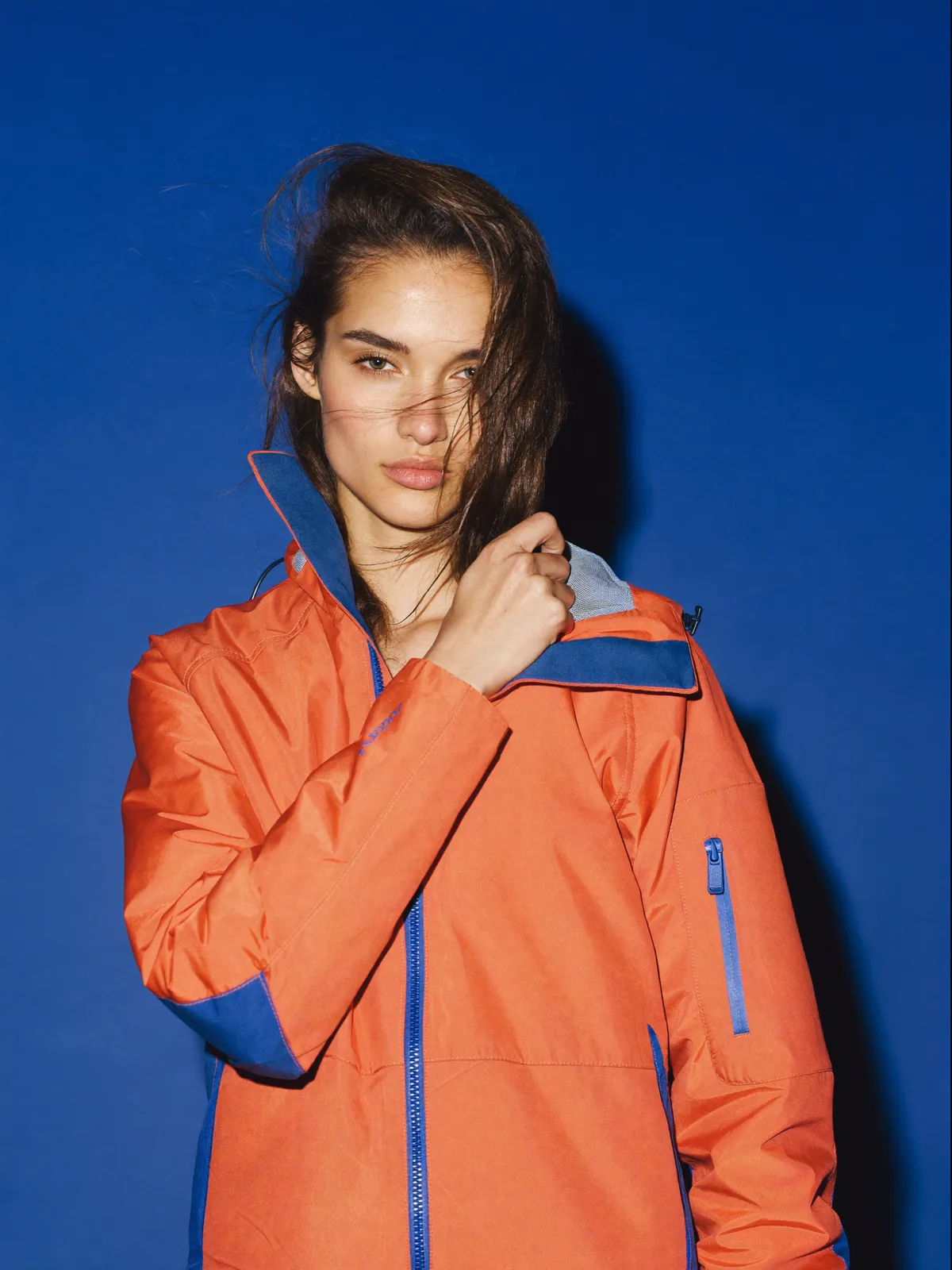 A fashion-editorial portrait of a model in an orange and cobalt-blue technical jacket against a deep blue backdrop, windswept hair across her face.