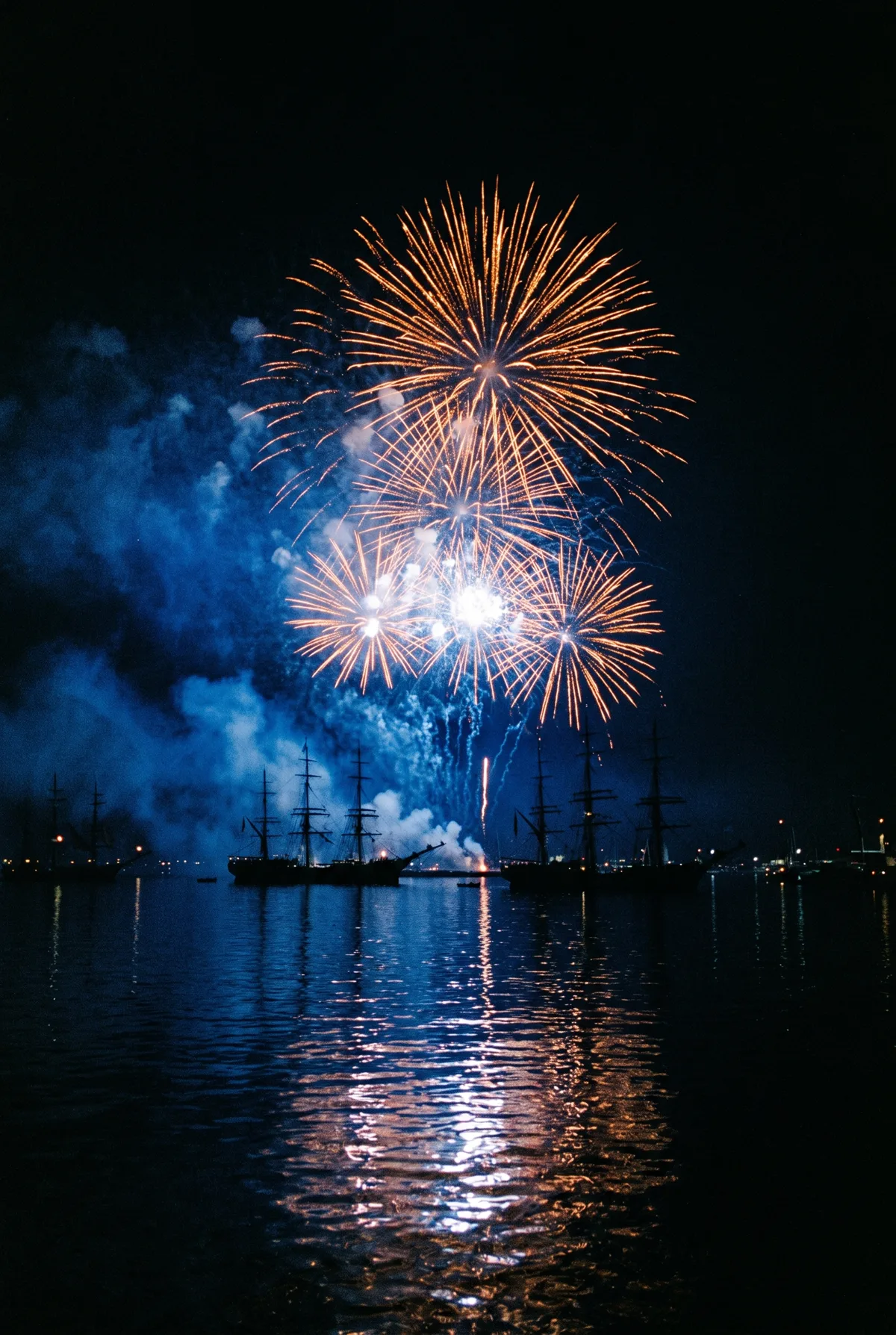 Orange and gold fireworks bursting over a night harbour, cobalt-blue gunsmoke drifting between silhouetted tall ships, reflections rippling in black water.